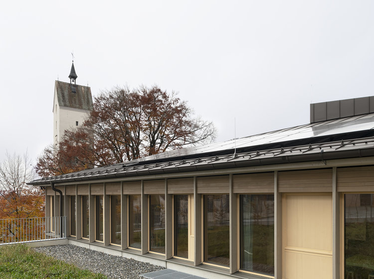 Modernes Flachdachgebäude mit großer Fensterfront und Holzfassade, umgeben von Grünfläche; im Hintergrund ein Kirchturm und herbstliche Bäume.